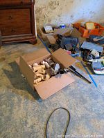 Multiple boxes on basement floor containing assorted plumbing fittings, drain cleaner tool, curtain rods, and other hardware.