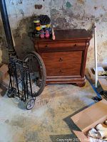 Photo of wooden cabinet showing three drawers with one drawer partially open and items on top. Loose knob visible. Cabinet placed in basement with stained walls and floor.