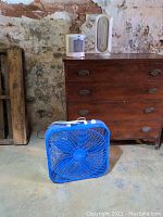 Blue Lasko box fan on floor with wooden chest background