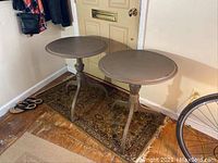 Pair of round wooden side tables shown next to a door on a patterned rug with bicycle wheel seen at edge of frame. One shows water stain.