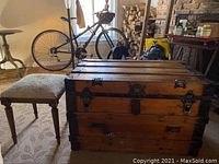 Photo showing the wooden trunk and upholstered foot stool placed side by side in a living room with other furniture and a bicycle in background.