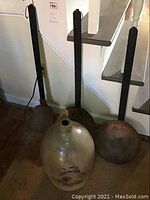 Full view of the large stoneware crock jug positioned on the floor with three long-handled iron cooking tools behind it, showing the general shape and condition of all items.