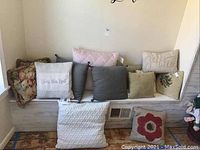 Thirteen assorted decorative throw pillows arranged on a white bench, showing a variety of colors and patterns including florals, stripes, and text designs.