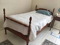 Full view of vintage wooden twin bed frame with white linens and blue patterned pillow on painted wood floor near side table.