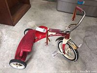 Side view of red Radio Flyer tricycle showing metal frame, red plastic rear platform, and white wheels with black tires. Visible rust spots and scratches.