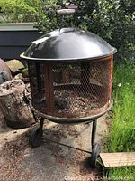 Full view of outdoor fire pit with rust on mesh sides, black domed lid with handle, and wheels for mobility.