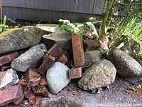 Large pile of irregular gray landscaping rocks mixed with several old red clay bricks, outdoors near a building.
