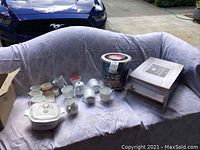 Full view of lot showing all items including the Corning Ware bakeware, multiple coffee cups, Christmas tin, and candles on a covered couch outdoors.