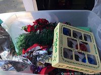 Assorted Christmas decorations inside a plastic bin: artificial greenery garland with red poinsettia flowers, silver reflective tin, string lights, red candle holder, and box of ornaments.