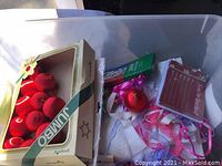 Photo of box with red plastic Christmas ornaments, pink and white ribbons, and invitation labels scattered in box.