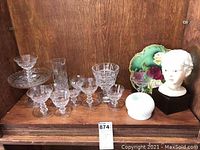 Multiple crystal and glass stemware, decorative floral plate, milk glass egg holder, coasters, and porcelain doll head bust arranged on wooden shelf.