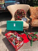 Christmas wreath placed on chair, green box, and Christmas tree stand with red base on the ground, box of assorted Christmas ornaments inside red container