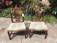 Two wooden chairs with carved backs and striped upholstered seats placed outdoors on a concrete surface with plants in the background.