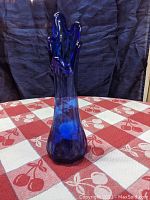 Front angle view showing rounded base and swung organic top edge of cobalt blue glass vase on checkered red and white cloth with dark background.