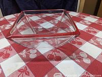 Side angle view showing the clear glass square bowl with prominently red rim on top placed on red and white checkered tablecloth with cherry patterns.