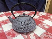 Full view of gray cast iron Japanese tea kettle with black handle and spout on a red and white tablecloth.