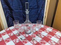 Full lot showing tall clear glass decanter with stopper and five matching wine stem glasses on a red and white checkered tablecloth.
