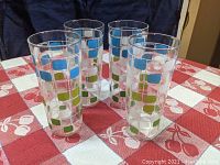 Four transparent glass tumblers standing upright on red and white checkered tablecloth with geometric square and rounded rectangle motif in blue, green, and white.