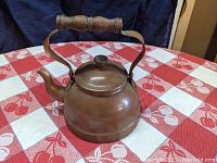 Copper tea kettle placed on a red and white tablecloth with cherry pattern, showing side view with wooden handle and spout.