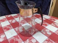 Clear glass Pyrex coffee carafe with black handle and gold starburst design on red and white checkered tablecloth, side view.