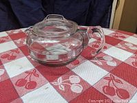 Side view of clear Pyrex glass tea kettle on red and white checkered tablecloth featuring a cherry pattern.