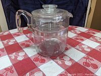 Clear glass Pyrex Flameware nine cup coffee percolator with handle, lid, and internal basket, photographed on a red and white checkered tablecloth.