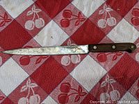 Full view of the 9-inch blade and wooden handle of the Comus utility knife lying on a red and white checkered cloth with cherry pattern.