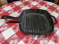Angled view of square cast iron grill skillet on red and white checkered tablecloth, shows long handle and well-defined ribbed grilling surface.