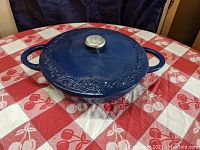 Front view of large blue enameled cast iron pan with lid on red and white checkered cloth. Shows two handles and lid with metal knob and decorative embossed design.