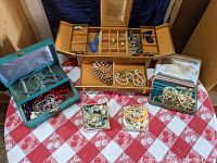Multiple boxes open showing assorted vintage costume jewelry including necklaces, earrings, and decorative pieces arranged on red and white checkered cloth.