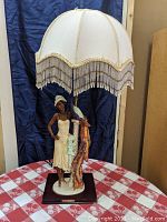 Full lamp with woman figurine, peacock, and fabric fringe lampshade on dark wooden base.