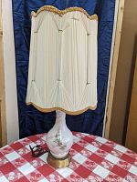 Full view of the lamp showing the white glass base with floral pattern, brass base, electrical cord, and drapery-style fabric shade with gold trim.