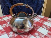 Front angle view of the rounded stainless steel tea kettle placed on a red and white tablecloth, showing the wood-toned handle and dome lid with knob.