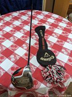 Full view of the left-handed golf club with graphite shaft and accompanying black knit head cover with pom-pom on a red and white checkered tablecloth.