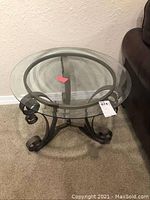 Glass top curved metal scrollwork base end table viewed diagonally from above and side showing glass top diameter and scroll foot detail.
