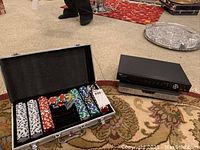 Poker set with chips in metal case next to two stacked electronic devices on carpet