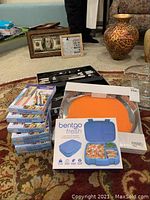 Photo showing seven boxed sets of 12-piece stainless steel tableware with pink handles, Bentgo Fresh lunch box, round orange metal serving tray, grilling tools in a case, and framed decorative items in the background.