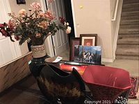 Photo displaying a large cream-colored vase with artificial flowers, black decorative crescent-shaped lamp base, and red fabric lamp shade with three framed wall art pieces near a staircase.