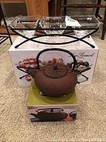 Full view showing the cast iron teapot on its box in front of a metal fruit bowl stand holding three glass bowls