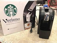 Starbucks Verismo pod brewer and original box displayed on carpeted floor with power cord visible.