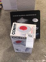 Conair infrared heat massager in box with red heating surface visible, placed in front of the luxury rainfall shower head box on beige carpet.