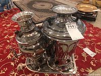 Metal samovar, urn, and tray set placed on a red patterned fabric on the floor with other household items in background.