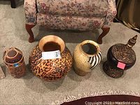 Four vases grouped on carpet, showing ceramic leopard print vase, cream vase with abstract design, dark glass vase with metal lid/base, and tall woven fabric covered vase