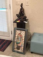 Full view showing pedestal with seated figure statue on top and metal wall art resting against it.