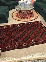 Two area rugs displayed on beige carpet floor. Rectangular rug with dark red, black, and white geometric pattern with fringe on edge. Round rug with cream center and red floral border curling at edges, placed near a glass coffee table.