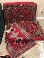 Photo of two large floor pillows with oriental pattern leaning against a wall, one pillow lying flat with a rug draped over it showing geometric designs in red and dark tones