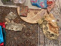 Overview of eight decorative fabric table covers and runners laid out on Persian rugs, showing different colors and styles mainly in gold tones with some blue and multicolor fabrics.