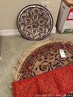 Photo shows the decorative copper plate with intricate symmetrical patterns. The round area rug with beige and red floral pattern is visible on the floor beneath. Part of the red fabric wall hanging is visible at the bottom of the image.