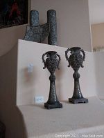 Two ornate candleholders displayed against a beige wall, showing height and detailed metalwork