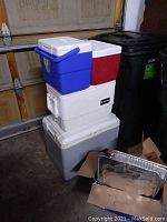 Stacked coolers in garage: blue, red, white, and gray coolers stacked vertically next to trash bins.